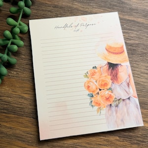 Puede incluir: Un bloc de notas rectangular con líneas horizontales y el texto Handfuls of Purpose impreso en la parte superior. El diseño presenta una ilustración de una figura con sombrero de paja que sostiene un ramo de rosas naranjas a la derecha.