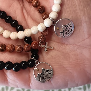 May include: Several beaded bracelets in shades of black, white, and brown. Some bracelets have a small cross charm or a round pendant with a mountain design and the text "Matt. 17:20". The beads appear to be made of stone or glass.