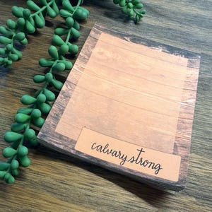 May include: A rectangular notepad featuring a printed wood grain pattern with horizontal planks. A light brown rectangular label at the bottom contains the black text "calvary strong" in a cursive script font. The notepad is positioned next to a green succulent.
