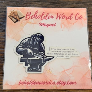 May include: A square magnet on a peach-coloured backing card featuring a black and white illustration of a blacksmith working at an anvil. The text reads: Iron sharpeneth iron: so a man sharpeneth the countenance of his friend. Proverbs 27:17.