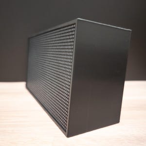 K&ouml;nnte beinhalten: Ein schwarzer rechteckiger Lautsprecher mit einer perforierten Frontplatte. Der Lautsprecher ist kastenf&ouml;rmig und steht auf einer hellen Oberfl&auml;che. Der Hintergrund ist dunkelgrau.