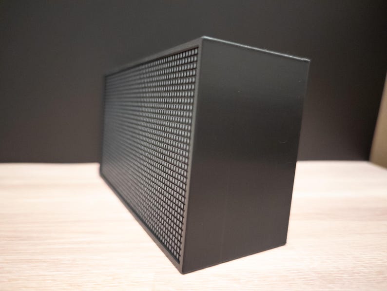 K&ouml;nnte beinhalten: Ein schwarzer rechteckiger Lautsprecher mit einer perforierten Frontplatte. Der Lautsprecher ist kastenf&ouml;rmig und steht auf einer hellen Oberfl&auml;che. Der Hintergrund ist dunkelgrau.