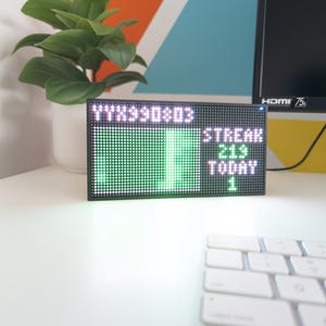 K&ouml;nnte beinhalten: Ein schwarzer LED-Bildschirm mit gr&uuml;nem und wei&szlig;em Text und Grafiken. Der Bildschirm zeigt die W&ouml;rter "STREAK 219 TODAY 1". Eine wei&szlig;e Tastatur befindet sich im Vordergrund. Eine Topfpflanze befindet sich im Hintergrund.