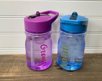 May include: Two clear plastic water bottles with colorful lids. One bottle has a purple lid and the name "Georgia" in pink. The other bottle has a blue lid and the name "Nolan" in green. Both bottles have a straw.