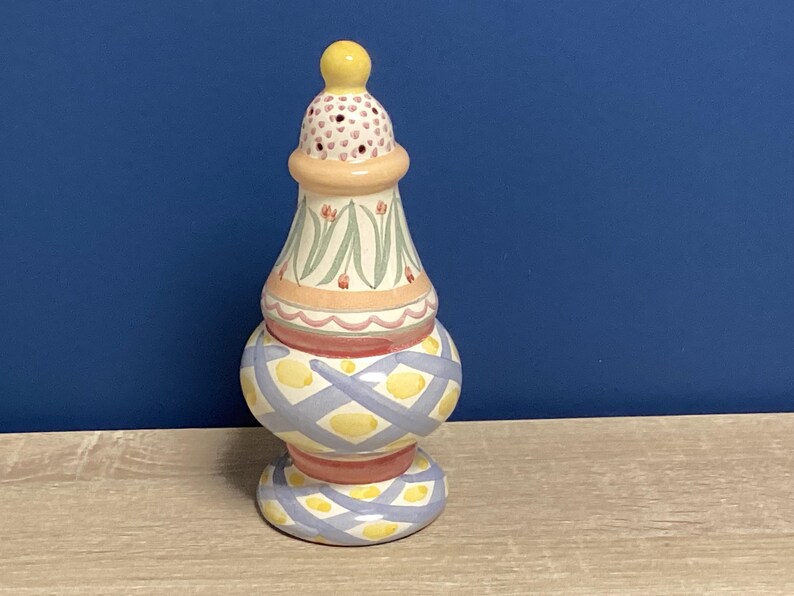May include: A decorative ceramic salt shaker with a yellow top and base. The body is painted with a light blue and yellow crisscross pattern. The middle section has a floral design with pink and green accents.