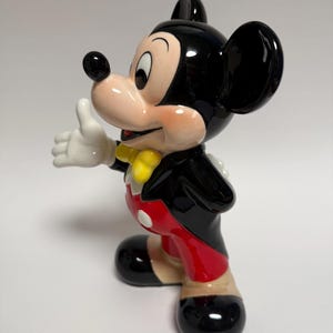 May include: A ceramic figurine of Mickey Mouse in a classic pose, with a thumbs-up gesture. The figure is predominantly black, with red shorts, yellow bow tie, and white gloves. The figurine is glossy and stands on a white surface.