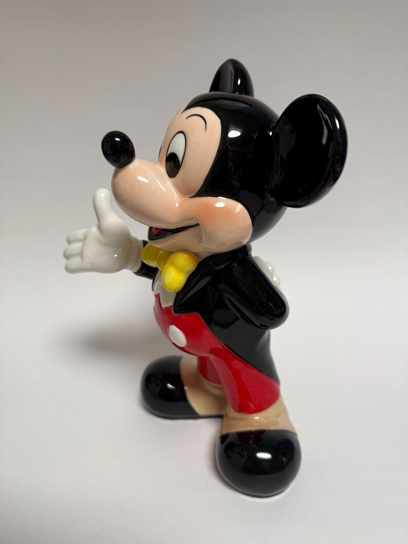May include: A ceramic figurine of Mickey Mouse in a classic pose, with a thumbs-up gesture. The figure is predominantly black, with red shorts, yellow bow tie, and white gloves. The figurine is glossy and stands on a white surface.