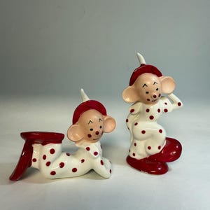 May include: Two ceramic mouse figurines. One is lying down, and the other is standing. Both are wearing white outfits with red polka dots and red hats and shoes. The figurines have tan faces with black eyes and noses.