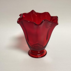 May include: A ruby red glass vase with a ruffled rim. The vase has a flared shape and a round base. The glass is transparent, allowing light to pass through. The vase is likely used for decorative purposes.