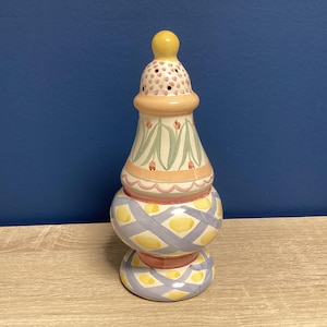 May include: A decorative ceramic salt shaker with a yellow top and a patterned design. The shaker features a bulbous base with a crisscross pattern in yellow and blue, and a floral design on the upper section.