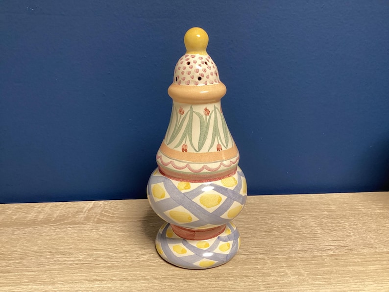 May include: A decorative ceramic salt shaker with a yellow top and a patterned design. The shaker features a bulbous base with a crisscross pattern in yellow and blue, and a floral design on the upper section.