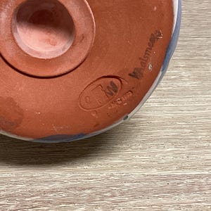May include: Close-up of the bottom of a ceramic object. The object is primarily reddish-brown with a circular opening and a raised rim. The object has blue and white accents and the text "Walsmere" is visible.