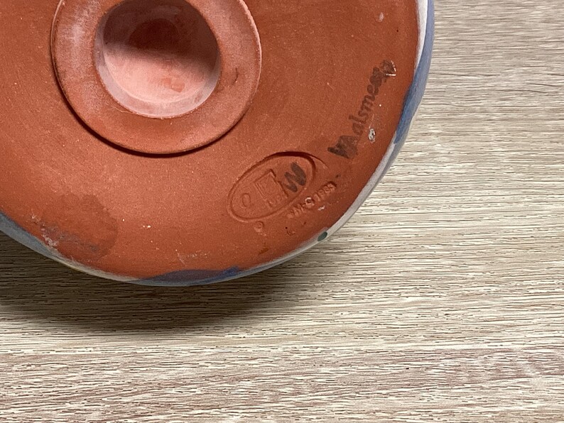 May include: Close-up of the bottom of a ceramic object. The object is primarily reddish-brown with a circular opening and a raised rim. The object has blue and white accents and the text "Walsmere" is visible.