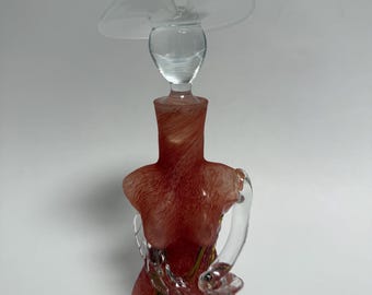 May include: A decorative glass sculpture in the shape of a woman, featuring a red-toned body and clear glass arms and hat. The sculpture has a swirling pattern and gold accents, creating an elegant and artistic design.