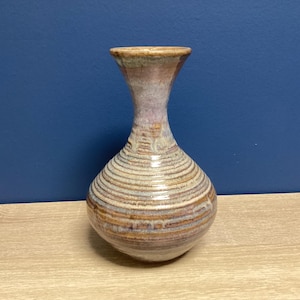 May include: A handcrafted ceramic vase with a bulbous base and a slender neck. The vase features horizontal, textured lines and a color palette of brown, tan, and purple. The vase sits on a wooden surface against a dark blue background.