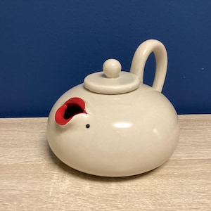 May include: A white ceramic teapot with a rounded body, a curved handle, and a small lid. The spout is a red, open design. A small black dot is on the side of the teapot. The teapot is on a light wood surface.