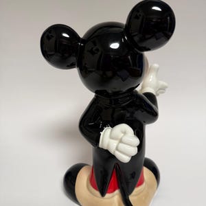 May include: A glossy black Mickey Mouse figurine with white gloves, red shorts, and tan shoes. The iconic character is posed with one hand raised and the other at his side. The figurine has large, round ears and a cheerful expression.