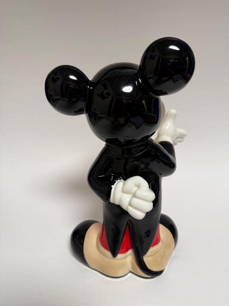 May include: A glossy black Mickey Mouse figurine with white gloves, red shorts, and tan shoes. The iconic character is posed with one hand raised and the other at his side. The figurine has large, round ears and a cheerful expression.