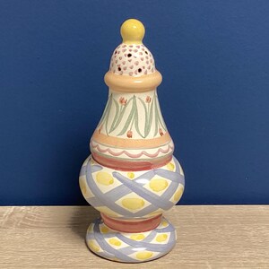 May include: A decorative ceramic salt shaker with a yellow knob on top. The shaker features a bulbous base with a blue and yellow diamond pattern, a central section with floral designs, and a pink and red trim.