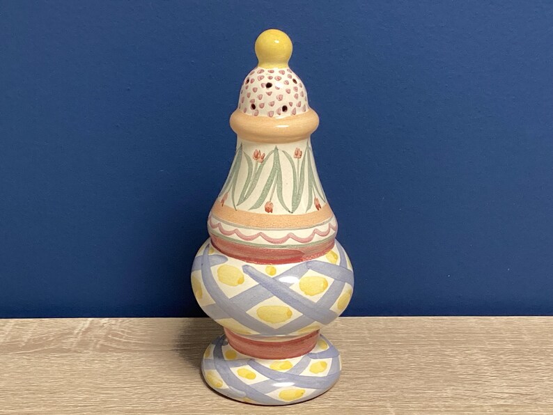 May include: A decorative ceramic salt shaker with a yellow knob on top. The shaker features a bulbous base with a blue and yellow diamond pattern, a central section with floral designs, and a pink and red trim.