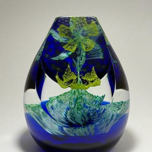 May include: A teardrop-shaped glass paperweight featuring a deep blue base and a clear upper section. Inside the glass, a vibrant green floral design with textured petals and a central yellow-green element is suspended, creating a layered effect.