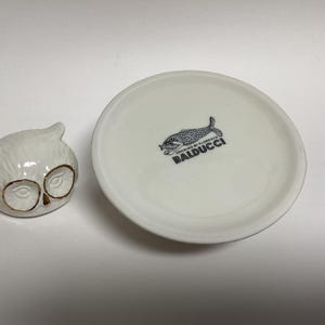 Puede incluir: Un recipiente de cer&aacute;mica blanca con forma de b&uacute;ho con detalles dorados en los ojos se encuentra junto a un plato blanco. El plato tiene una ilustraci&oacute;n de pez en blanco y negro y el texto "BALDUCCI".