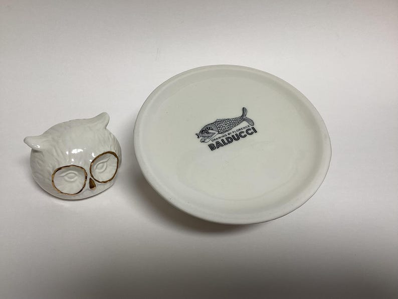 Puede incluir: Un recipiente de cer&aacute;mica blanca con forma de b&uacute;ho con detalles dorados en los ojos se encuentra junto a un plato blanco. El plato tiene una ilustraci&oacute;n de pez en blanco y negro y el texto "BALDUCCI".