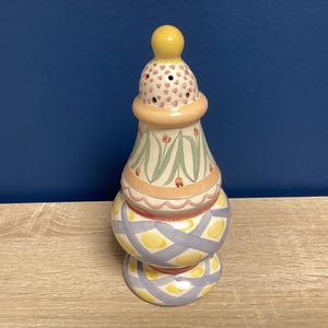May include: A decorative ceramic candle holder with a yellow knob on top. The candle holder has a bulbous base and a patterned design with pastel colors, including yellow, pink, and blue. The top section has small holes.
