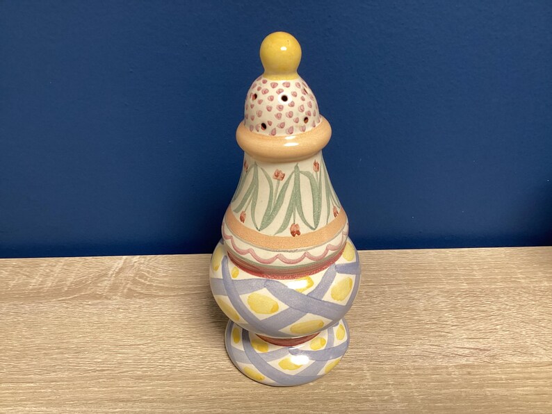 May include: A decorative ceramic candle holder with a yellow knob on top. The candle holder has a bulbous base and a patterned design with pastel colors, including yellow, pink, and blue. The top section has small holes.