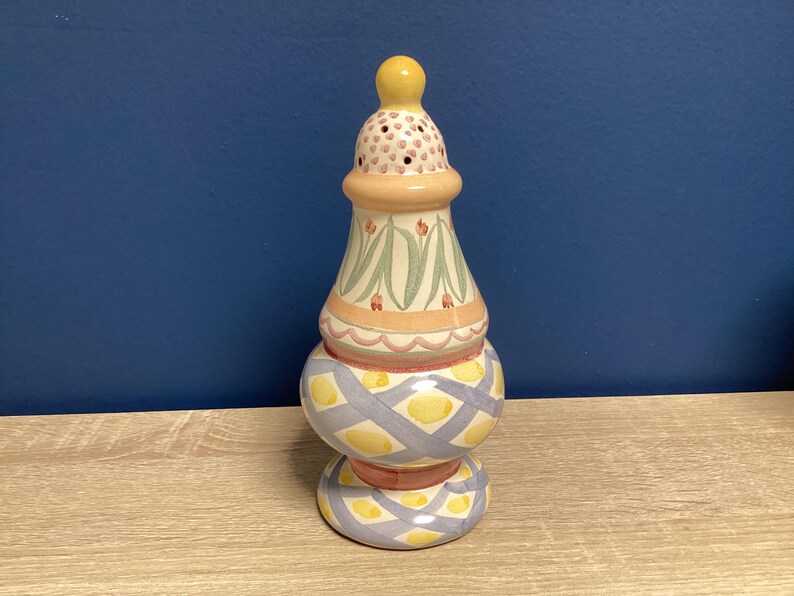 May include: A decorative ceramic salt shaker with a yellow, blue, and peach color scheme. The top has a yellow finial and a perforated section. The middle section features a floral design, and the base has a geometric pattern.