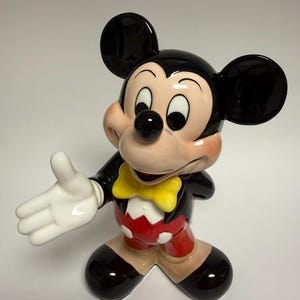 May include: A ceramic Mickey Mouse figurine. The figure has a black head and ears, a tan face, and a black nose. It wears a black jacket, red shorts, and a yellow bow tie. The figurine is posed with one arm outstretched.