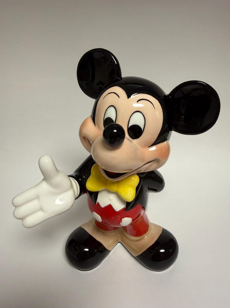 May include: A ceramic Mickey Mouse figurine. The figure has a black head and ears, a tan face, and a black nose. It wears a black jacket, red shorts, and a yellow bow tie. The figurine is posed with one arm outstretched.