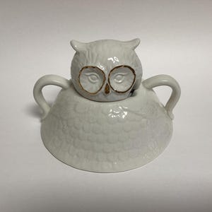 Puede incluir: Un recipiente blanco de cer&aacute;mica con forma de b&uacute;ho y tapa. El b&uacute;ho tiene gafas con borde dorado y un cuerpo texturizado. El recipiente tiene dos asas y probablemente se utiliza para servir o guardar.