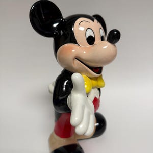May include: A ceramic Mickey Mouse figurine. The figure is in a classic pose, wearing a black jacket, red shorts, and a yellow bow tie. The figurine has black ears, a pink face, and white gloves. The figure is smiling and has one hand raised.