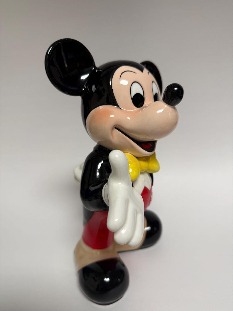May include: A ceramic Mickey Mouse figurine. The figure is in a classic pose, wearing a black jacket, red shorts, and a yellow bow tie. The figurine has black ears, a pink face, and white gloves. The figure is smiling and has one hand raised.