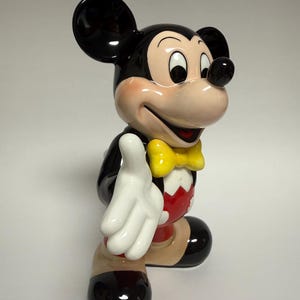 May include: A ceramic Mickey Mouse figurine, featuring a black body, large ears, and a smiling face with rosy cheeks. He wears a yellow bow tie, red shorts, and white gloves, with a friendly pose.