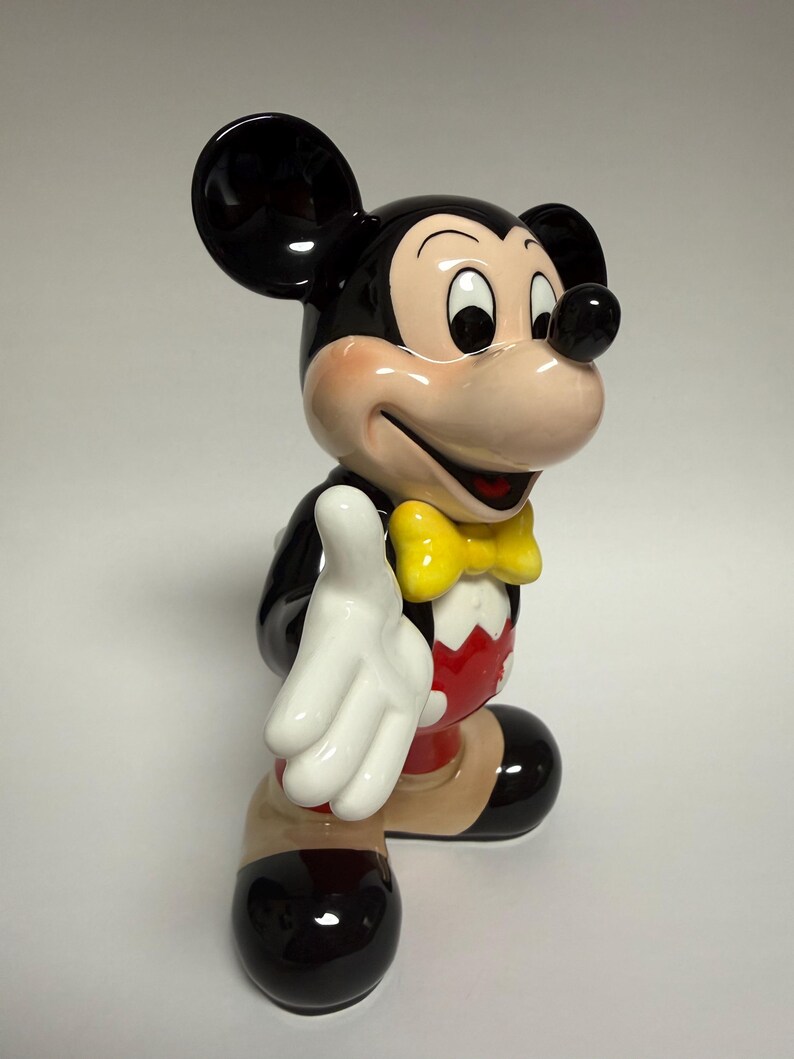 May include: A ceramic Mickey Mouse figurine, featuring a black body, large ears, and a smiling face with rosy cheeks. He wears a yellow bow tie, red shorts, and white gloves, with a friendly pose.
