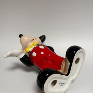 May include: A ceramic Mickey Mouse figurine, lying on its back. The figure has a black head and ears, a tan face, white gloves, a yellow bow tie, and red shorts with white polka dots. The base is white with the text "&copy;DISNEY JAPAN".