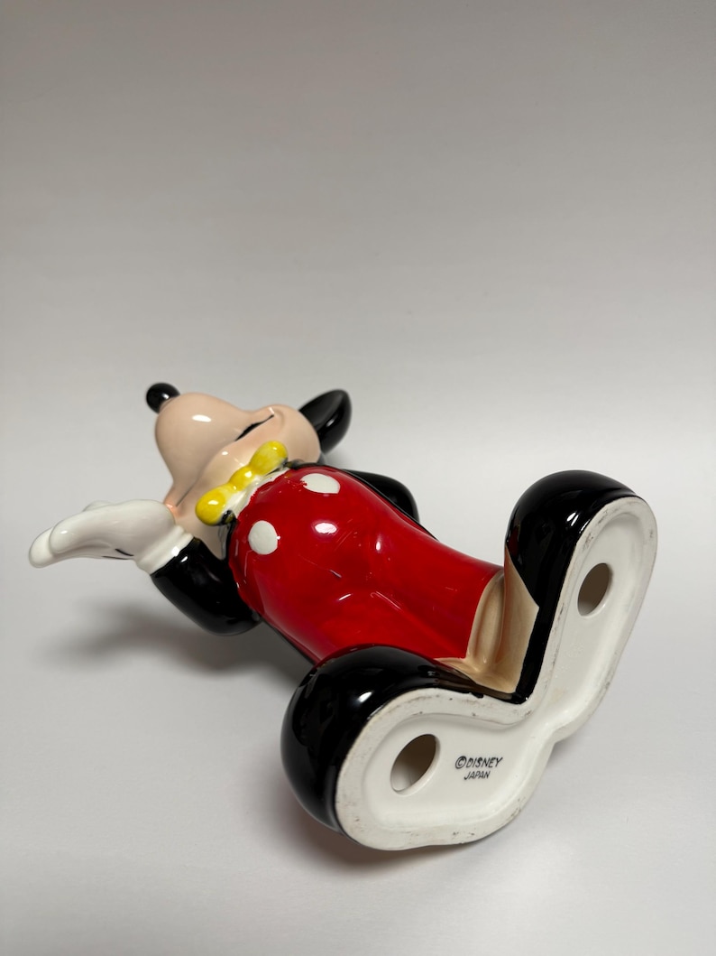May include: A ceramic Mickey Mouse figurine, lying on its back. The figure has a black head and ears, a tan face, white gloves, a yellow bow tie, and red shorts with white polka dots. The base is white with the text "&copy;DISNEY JAPAN".