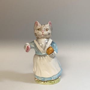 May include: A ceramic figurine of a cat dressed in a blue and white outfit, holding a bar of soap and a golden object. The cat has gray fur with pink accents on its ears. The word "SOAP" is visible on the soap.