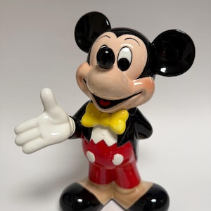 May include: A ceramic Mickey Mouse figurine in a classic pose. Mickey is wearing a black jacket, red shorts, and a yellow bow tie. His large black ears and white gloves are prominent. The figurine has a glossy finish.