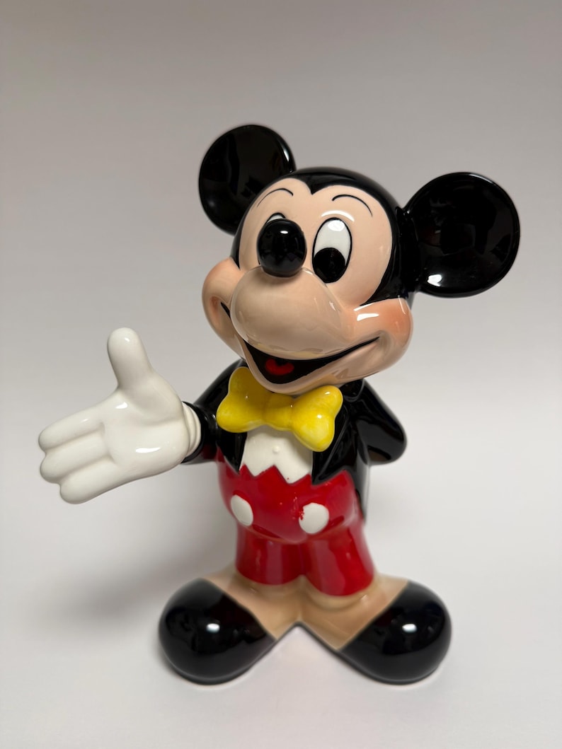 May include: A ceramic Mickey Mouse figurine in a classic pose. Mickey is wearing a black jacket, red shorts, and a yellow bow tie. His large black ears and white gloves are prominent. The figurine has a glossy finish.