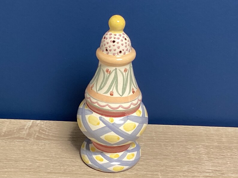 May include: A decorative ceramic vase with a yellow finial. The vase features a stacked design with painted floral and geometric patterns in pastel colors, including pink, blue, and yellow. The vase is approximately 8 inches tall.
