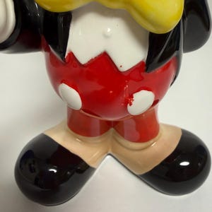 May include: A ceramic figurine of a cartoon character, featuring a black jacket, red shorts, and black shoes. The character has a yellow bow tie and white gloves. The figurine is glossy and has a classic design.