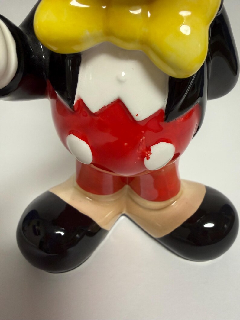 May include: A ceramic figurine of a cartoon character, featuring a black jacket, red shorts, and black shoes. The character has a yellow bow tie and white gloves. The figurine is glossy and has a classic design.