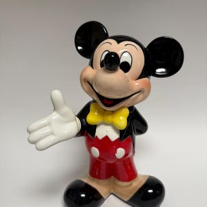 May include: A ceramic Mickey Mouse figurine. The figure is wearing a black jacket, red shorts with white buttons, a yellow bow tie, and black shoes. Mickey has a smiling expression and is extending his right hand.