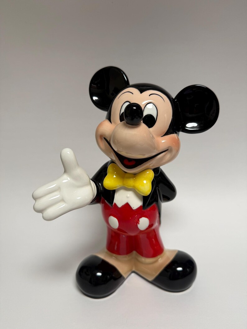 May include: A ceramic Mickey Mouse figurine. The figure is wearing a black jacket, red shorts with white buttons, a yellow bow tie, and black shoes. Mickey has a smiling expression and is extending his right hand.
