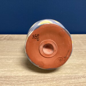 May include: A ceramic, cylindrical object with a rounded base. The exterior features a blue and white pattern with yellow accents. The base is reddish-brown with a central hole. The number "360" is written on the side.