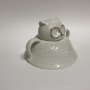 Puede incluir: Un recipiente de cer&aacute;mica blanco con forma de b&uacute;ho con tapa. El b&uacute;ho tiene detalles dorados alrededor de los ojos. El recipiente tiene un dise&ntilde;o texturizado de plumas y dos asas curvas. La tapa tiene forma de cabeza de b&uacute;ho.