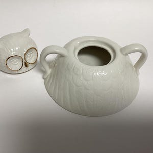 Puede incluir: Un azucarero de cer&aacute;mica blanca con forma de b&uacute;ho con tapa a juego. La tapa es una peque&ntilde;a cabeza de b&uacute;ho con detalles dorados en los ojos. El cuenco tiene dos asas y un dise&ntilde;o de plumas texturizadas.